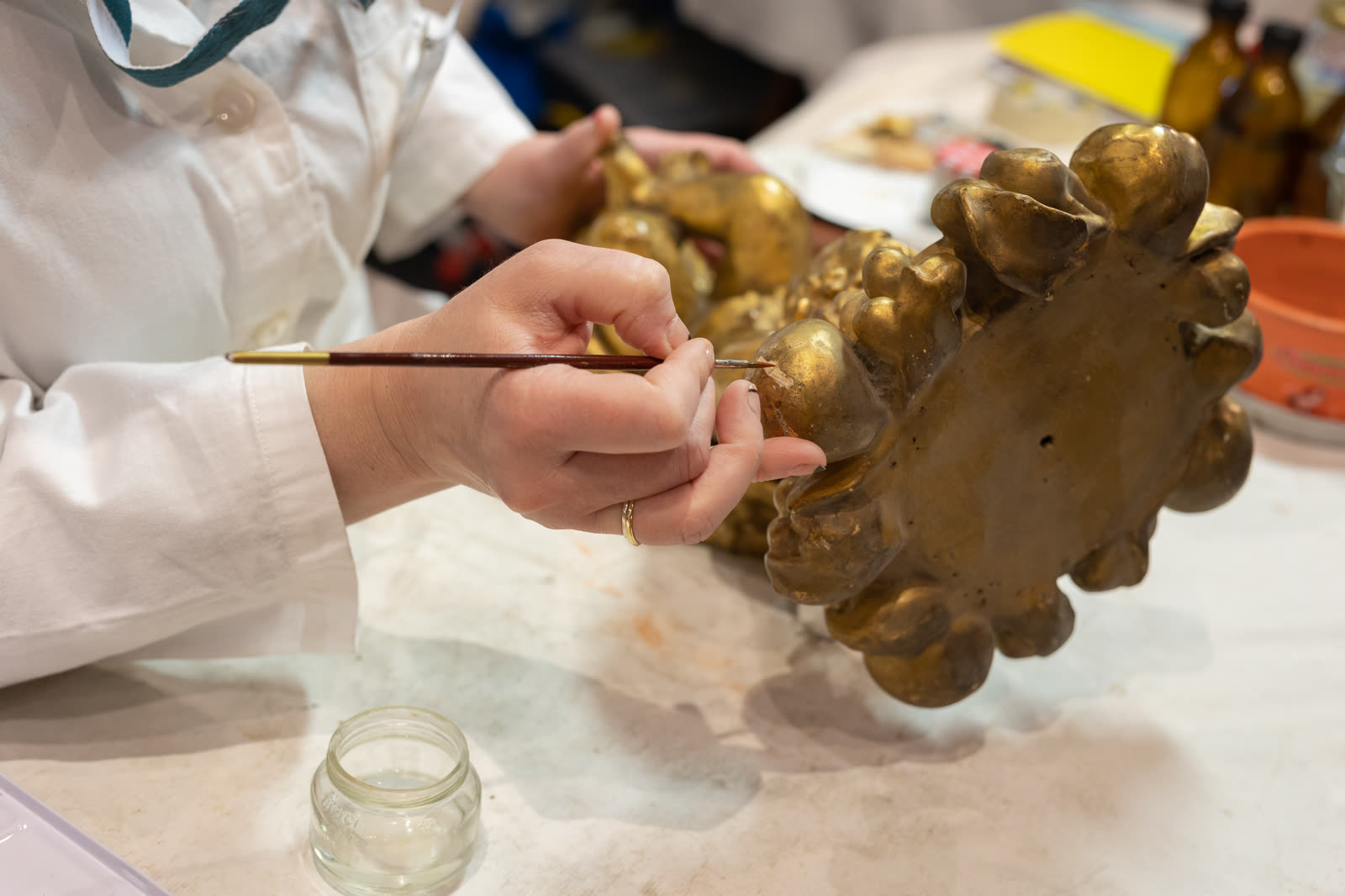 A hand in a white smock applies gold pigment with a fine brush to the textured surface of an ornate gilded bronze statuette.