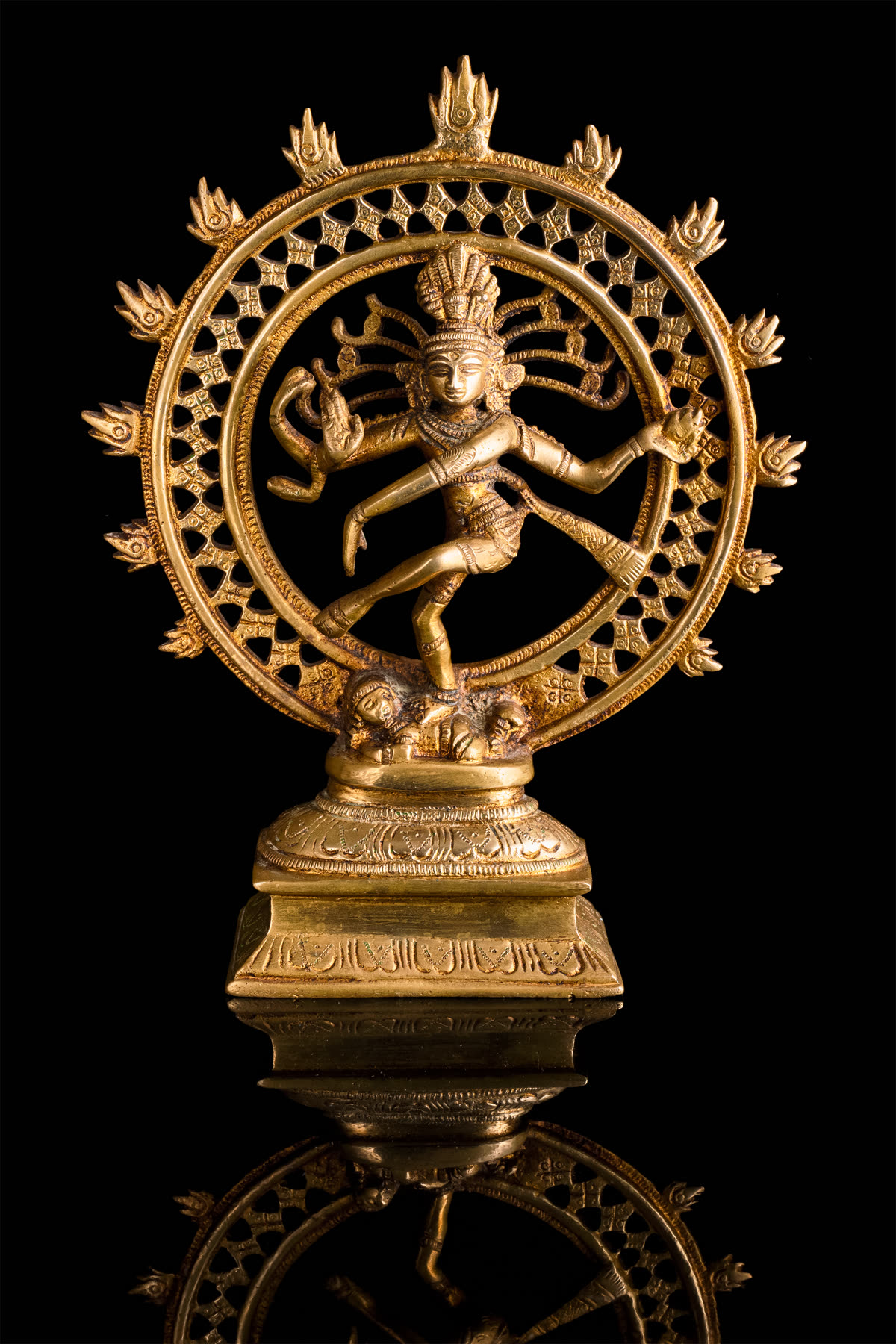 A gilt-bronze Nataraja statuette, Shiva dancing within a flaming halo ring on a carved pedestal, photographed against black.