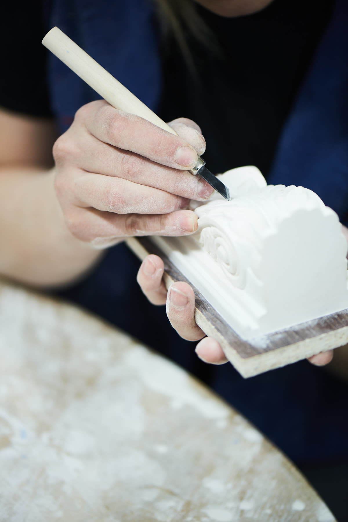 A craftsperson's plaster-dusted hand carves fine scrollwork detail into a pale plaster relief with a small chisel.