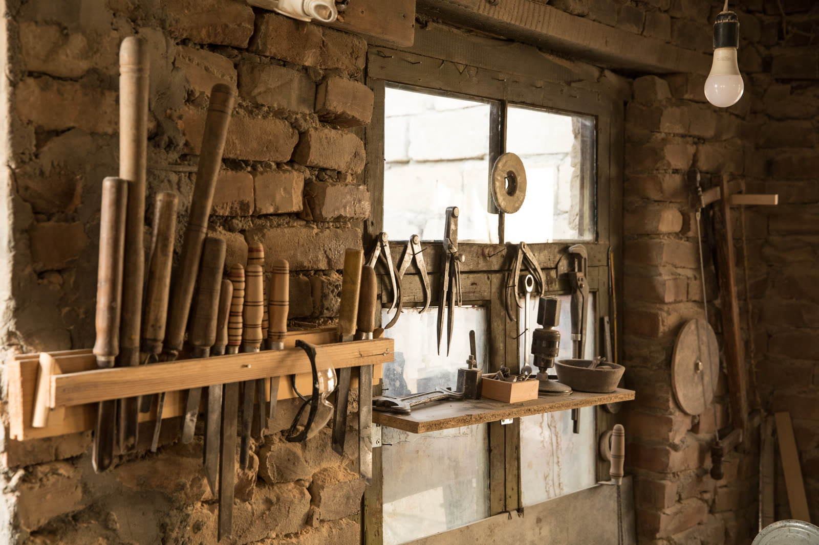 Rows of wood-handled chisels and files hang from a rack on a rough brick wall beside a window in a stone-walled workshop.